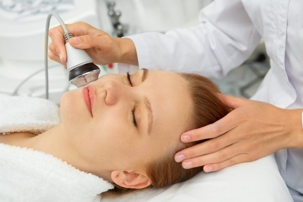 Beautiful young woman smiling while receiving facial ultrasonic skincare treatment by professional cosmetologist wellbeing pampering dermatology fresh smooth recreation spa salon professionalism